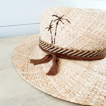 St. Barts raffia hat made of natural raffia. Crown of hat is finished with palm tree pyrography and wrapped with a striped jute band and a sepia gauze tie. The other side has a piece of Palo Santo wood hand stitched to the side. Hand crafted by La Macarena Hats in Tulum Mexico.