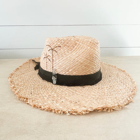 Boheme Raffia Hat with black suede and cotton gauze strips wrapped around the crown. Finished with palm tree pyrography on the side and a hand sewn white quartz crystal. Made of natural raffia with raw edges on the brim. Artisan made my La Macarena Hats in Tulum Mexico.