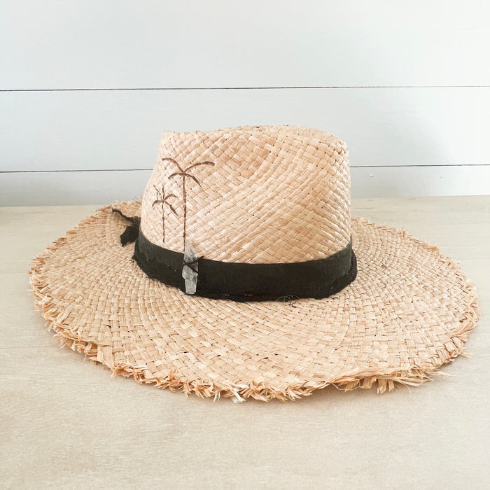 Boheme Raffia Hat with black suede and cotton gauze strips wrapped around the crown. Finished with palm tree pyrography on the side and a hand sewn white quartz crystal. Made of natural raffia with raw edges on the brim. Artisan made my La Macarena Hats in Tulum Mexico.