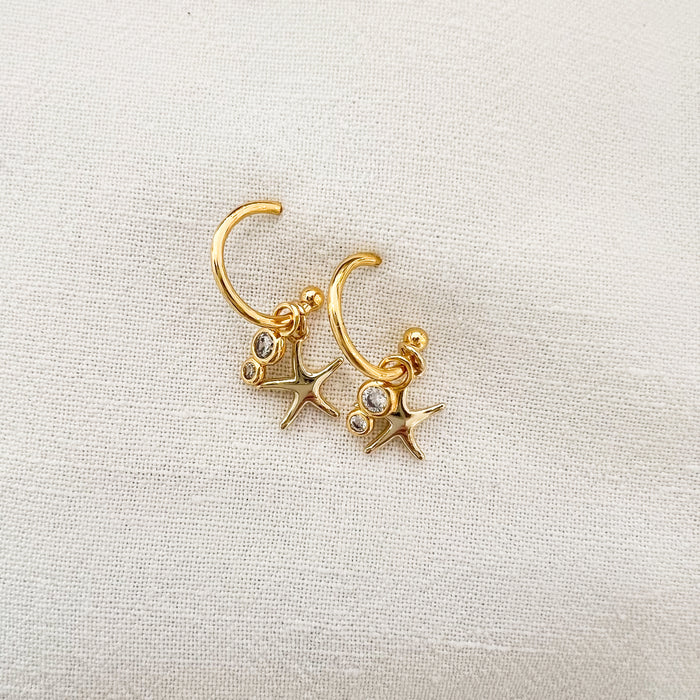 Pair of delicate open ended gold  hoop earrings with a mini gold starfish charm and a tiny double CZ crystal drop charm.  18k gold plated over brass with e-coating to protect the finish. .5" diameter 1" length.