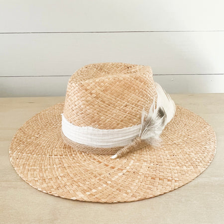 Walk-About hat woven in natural raffia. Crown is wrapped in a white cotton gauze strip and a natural braided jute strap. A bouquet of white and brown feathers are stitched to the side. Made by La Macarena Hats.