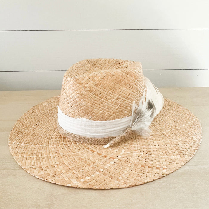 Walk-About hat woven in natural raffia. Crown is wrapped in a white cotton gauze strip and a natural braided jute strap. A bouquet of white and brown feathers are stitched to the side. Made by La Macarena Hats.