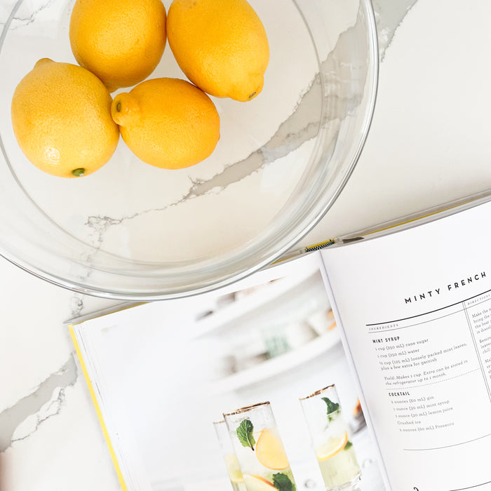 Aero glass serving bowl holding yellow lemons next to an open cookbook. Lemons and cookbook not included. Clear glass bowl in a modern curved concave silhouette. Measures 11.75" x 4.25".