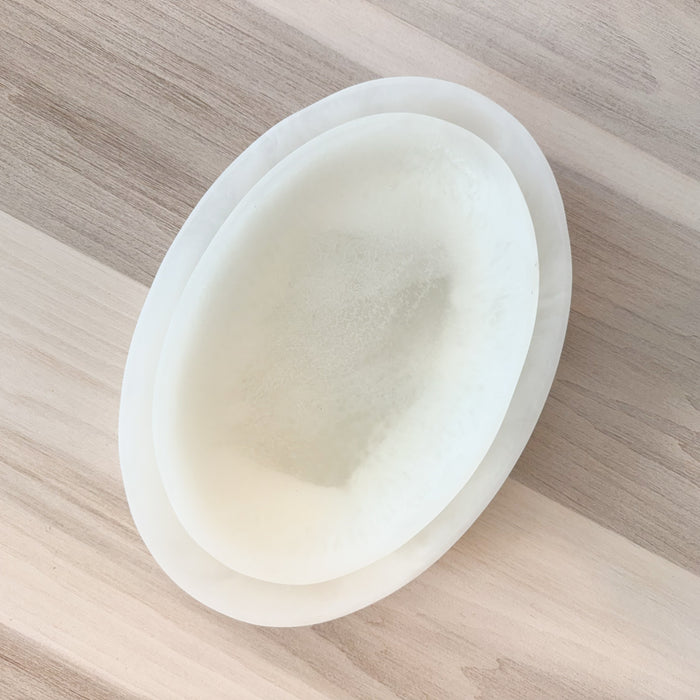 Stack of two oval shaped white resin platters on a pale wood surface. Medium 11.5 x 7 inches. Large 14 x 10 inches. Each sold separately.
