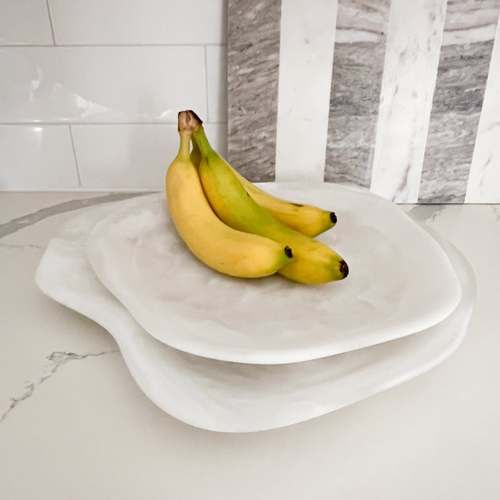 Amalfi serving platters styled on kitchen counter with bananas. Two white serving platters in a curvy organic shape crafted from swirls of white opaque and translucent resin. Luxury resin serving pieces suitable for indoor/outdoor entertaining. Each sold separately.