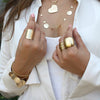 Close up shot of model wearing white shirt and hands up wearing gold rings featuring the Vionna Ring, the Cluster Wire Wrap Ring and the Lorabelle Dome Ring. Each sold separately.