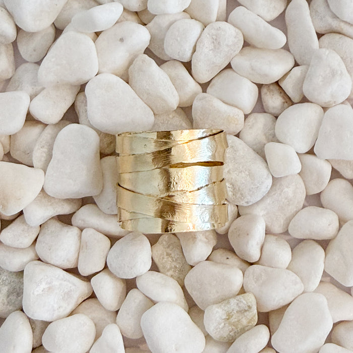A textured gold ring with a multi-spiral, layered design, resting on a bed of smooth white pebbles. 18k gold over brass. Adjustable, fits size 7-9.