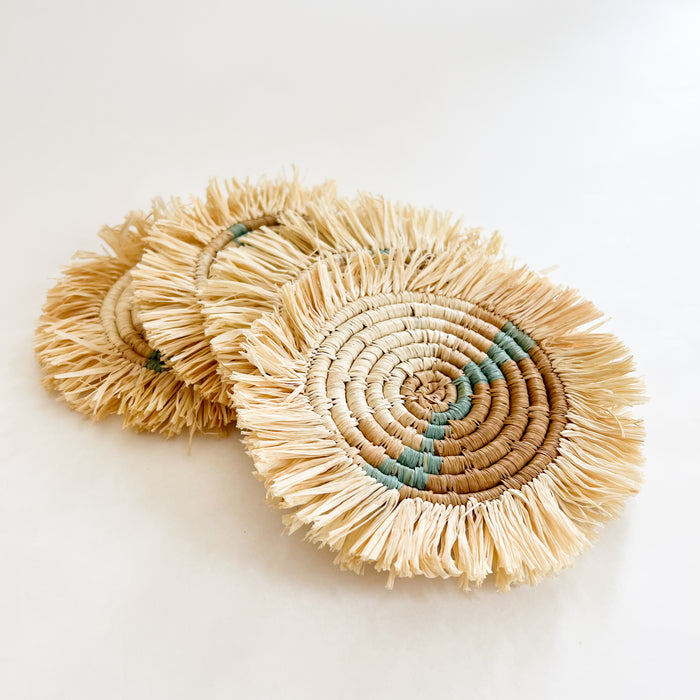 Set of 4 Dunes Seagrass coasters. Round coasters handwoven in  an abstract pattern using natural seagrass, sepia and sea green. Finished with a natural raffia fringe trim. Measures 6” diameter. Fair trade, made by female artisans in Africa.