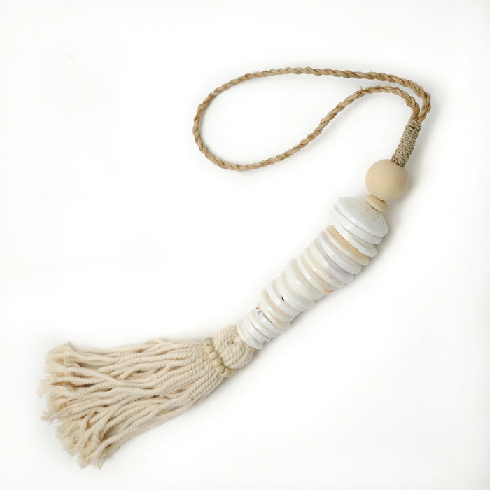A bohemian-style tassel ornament featuring a cream cotton tassel, stacked white shell discs, a natural wood bead, and a braided jute loop on a white background. Measures 26" length. Hand crafted by artisans in Indonesia.