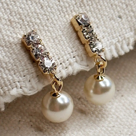 Pair of triple CZ stone stud earrings with a white pearl drop on a natural canvas  ground. 18K gold filled, simulated pearl. Water resistant.