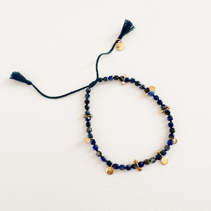 Lapis and gold corded bracelet. Square cut Lapis stones interspersed with gold filled charms on an indigo cord. Adjustable tassel pulls. Hand crafted by TAI Jewelry.