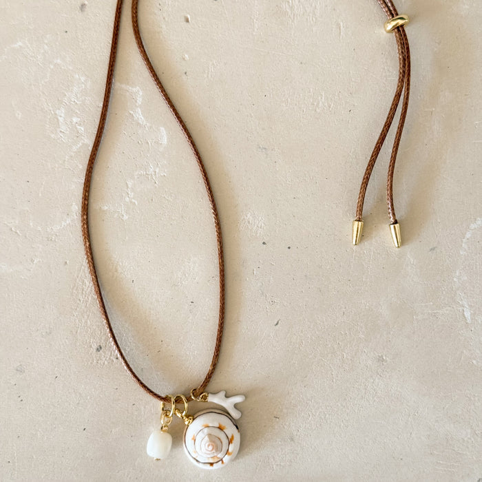 A bohemian-style necklace featuring a tan waxed bolo cord, a white pearl, a white spiral seashell pendant, and a small faux white coral fragment. 26" length with 18k gold filled adjuster and tips.
