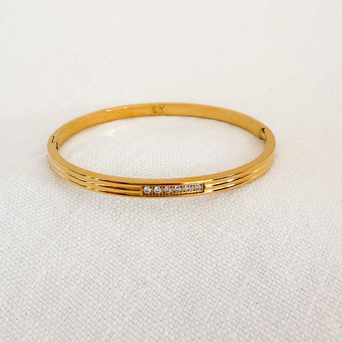 Delicate oval shaped gold fluted bangle with a row of five sparkling clear CZ stones. Hinged closure. Made of marine grade stainless steel with gold plating and special protective coating. Water, sweat and tarnish resistant.