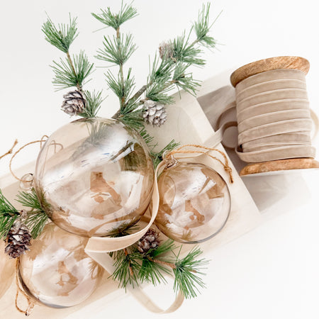 Taupe tinted glass ball ornaments with taupe velvet ribbon and evergreens styled in a bleached wood tray. Each ornament sold separately.