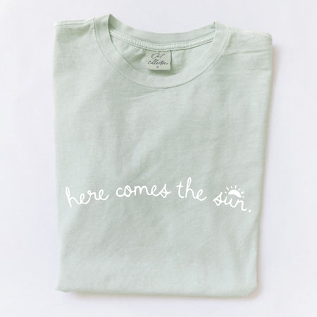 Folded light green t-shirt with "here comes the sun" text on a white background.