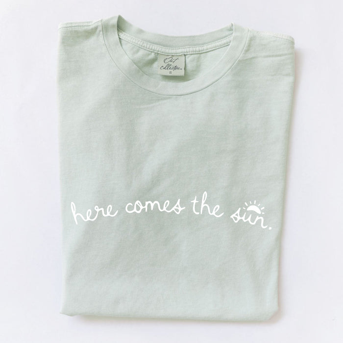 Folded light green t-shirt with "here comes the sun" text on a white background.