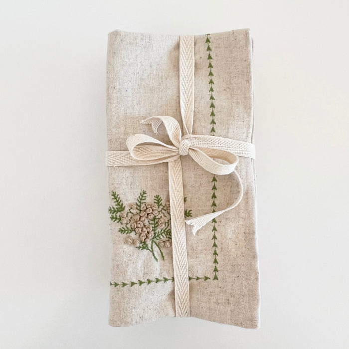 Neutral linen blend napkins with a pine green embroidered border feature embroidered pine branches with tiny brown french knot pinecones in one corner. Set of 4 18" square napkins come bundled in natural twill tape.