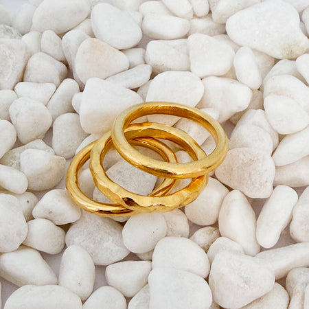 Three stacked gold rings one with a smooth brushed finish, one with a hammered finish and one with a polished finish laying on white pebbles. 18k gold over brass.