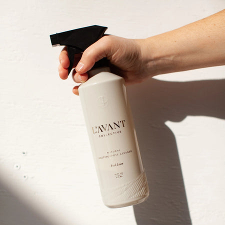 Hand holding a beige spray bottle with black nozzle. Multipurpose spray cleaner by L'AVANT in fresh linen scent. 16 fl oz./475 ml.