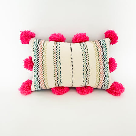 Decorative lumbar pillow made from a colorful Mexican textile. Multi colored stripes on an ecru background  trimmed with hot pink pompoms on all four sides. 17 x 11 inches.