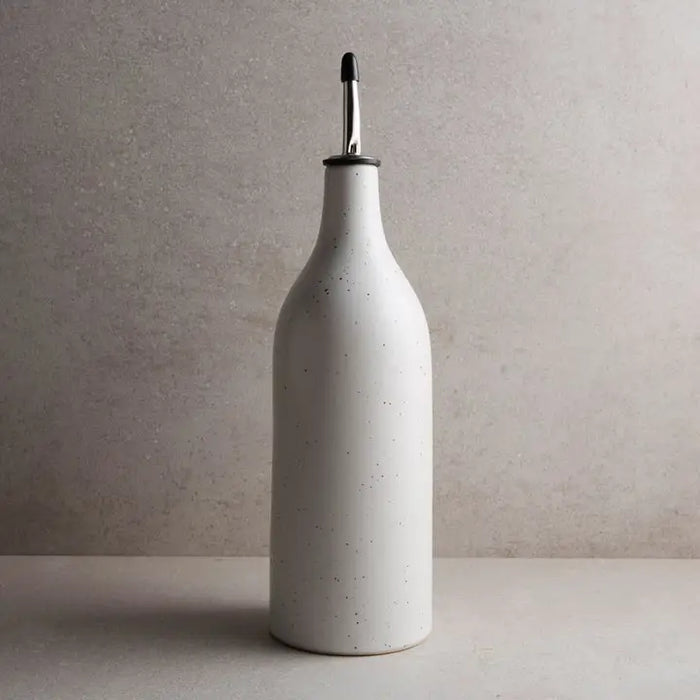 A large white stoneware oil cruet with a speckled glaze and silver metal spout sitting on a beige counter. 12 inch tall, 3.25 inch diameter.