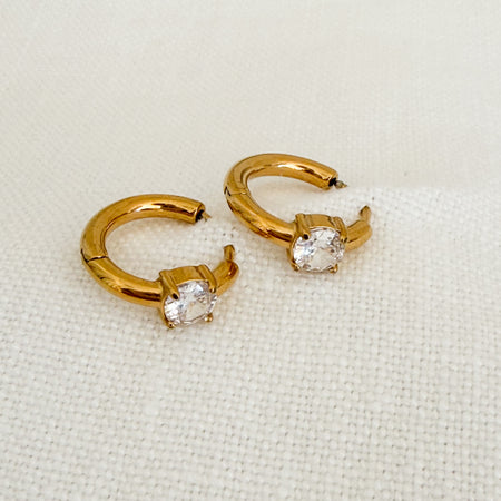 Pair of small gold huggie hoop earrings with a clear solitaire CZ stone. Clicker back. Water, sweat and tarnish resistant. Made of hypoallergenic stainless steel with gold plating.