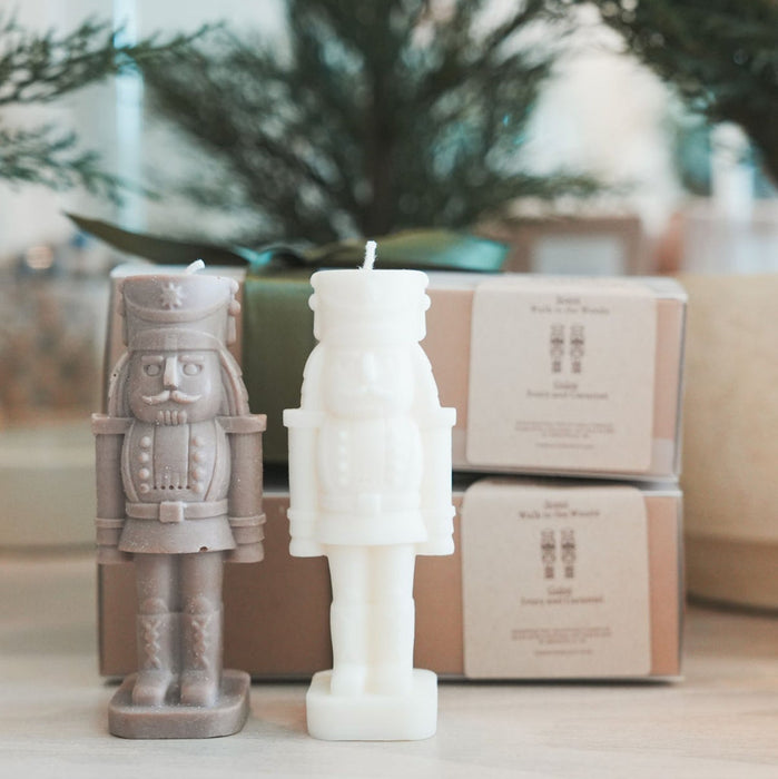 Two nutcracker-shaped candles. One warm taupe, the other in ivory. "Walk in the Woods" fragrance has the scent of fresh evergreens. Comes in a Kraft gift box wit a green satin bow.
