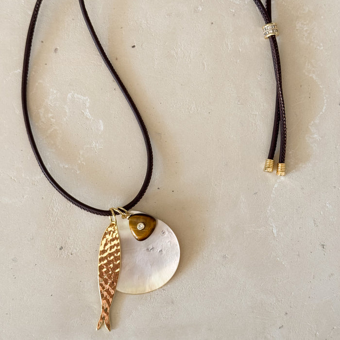 A brown leather bolo cord necklace with three pendants: a golden hammered fish, an abstract white shell disc and a Tigers Eye stone embedded with a clear CZ stone. 18" length with pave' slide adjuster.
