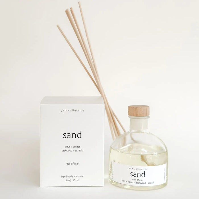 SAND reed fragrance diffuser in clear bottle with natural rattan reeds next to a white box with SAND by yam collective printed on it. Fragrance notes of Citrus, Amber, Teakwood and Sea Salt. 5 fl oz.