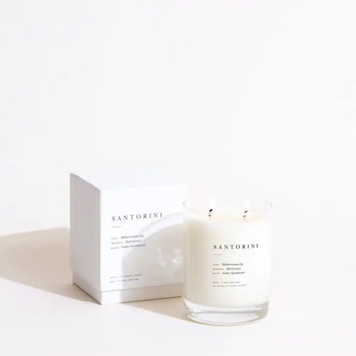 Santorini Candle. Luxe scented soy wax candle in clear glass vessel. Packaged in a matte ivory box with minimalistic black typography. Fragrance base notes of Sandalwood and Amber, mid notes of Red Currant and top note of Mediterranean Fig. Approximate burn time 70 hours. 13 oz. volume