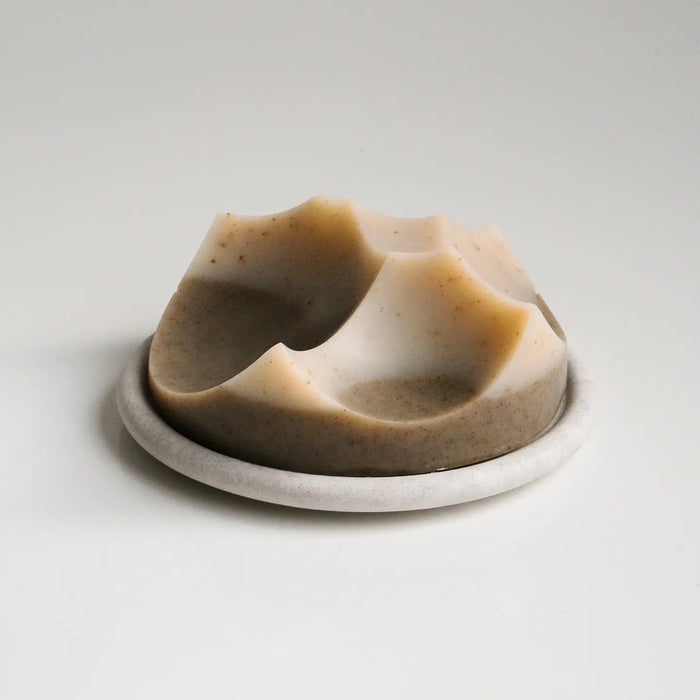 Tan and cream sculpted soap bar on a white soap dish on a grey background. Sculpted Cold Press soap in Cardamom Vetiver fragrance by Erode.