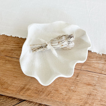 White ceramic seashell dish/catchall styled with a smudge stick (not included). Shell shaped catchall ceramic dish with a sandy white glaze. Perfect as a jewelry dish or pretty catchall for the coastal home. 7.75 x 6.75 inches.