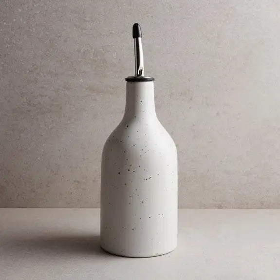 Small white stoneware oil cruet with a speckled glaze and silver metal spout sitting on a beige counter. 9 inches tall, 3 inch diameter.