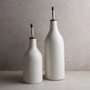 Two white stoneware oil cruets with speckled glaze and silver metal spouts. One small and one large on a taupe counter. Each sold separately.
