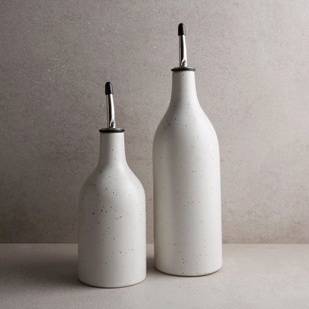 Two white stoneware oil cruets with speckled glaze and silver metal spouts. One small and one large on a taupe counter. Each sold separately.