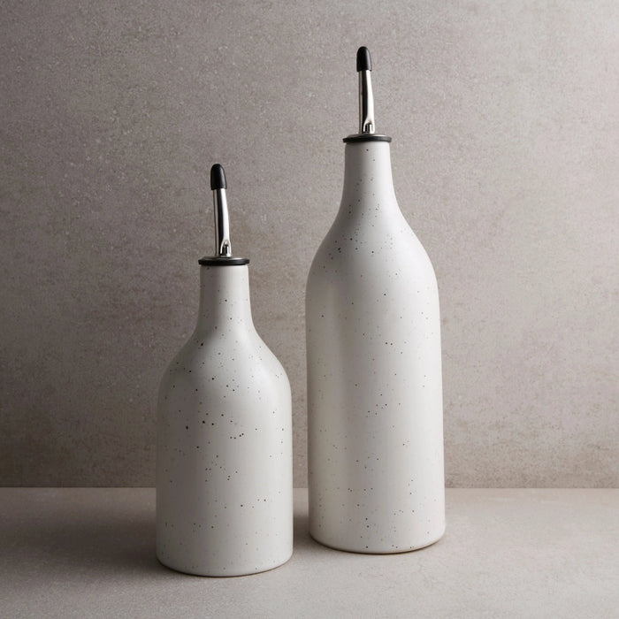 Two white stoneware oil cruets with speckled glaze and silver metal spouts. One small and one large on a taupe counter. Each sold separately.