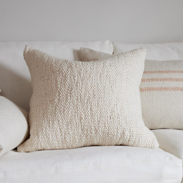 A square ivory throw pillow with a textured boucle surface, styled on a white sofa with other neutral-toned pillows in the background. Each pillow sold separately.