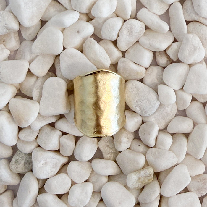 A bold hammered gold ring with a wide, open band, resting on a bed of small white pebbles. Adjustable, fits size 7-9. 18k gold over brass.