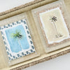 Two watercolor painting of palm trees, on in shades of blue the other in sandy neutrals. Each framed with a tonal decorative woodblock print matte in a clear lucite block. Displayed on a seagrass tray.