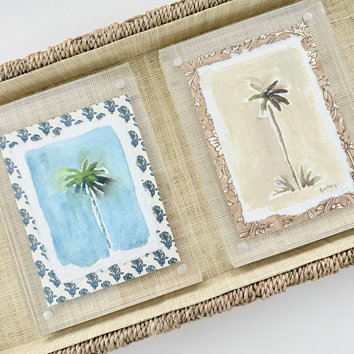 Two watercolor painting of palm trees, on in shades of blue the other in sandy neutrals. Each framed with a tonal decorative woodblock print matte in a clear lucite block. Displayed on a seagrass tray.