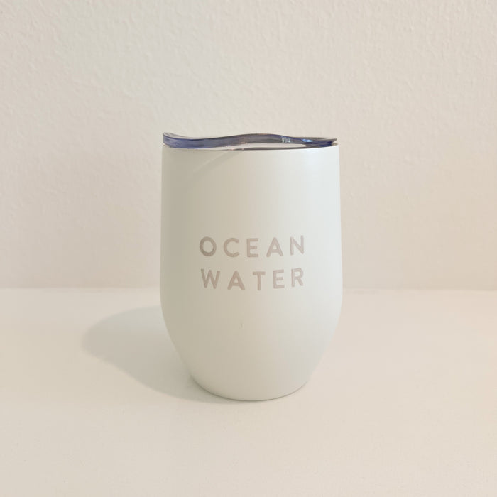 Ocean Water, stainless steel stemless wine tumbler with clear  sip lid. Matte white enamel exterior with glossy cream lettering "OCEAN WATER". Holds 12 fl ounces.