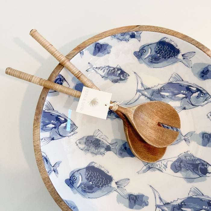 Pesce Serving Bowl. Natural mango wood bowl with a white food safe resin interior featuring a blue watercolor fish design. Measures 12.75" diameter 3" height. Shown with wood + rattan salad servers, sold separately.