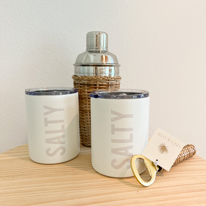 SALTY stainless steel tumblers shown with rattan cocktail shaker and rattan handled bottle opening. Each sold separately. Insulated stainless steel tumbler with clear sealed sip lid. Exterior is matte white enamel with glossy cream lettering "SALTY". Holds 12 fl ounces.