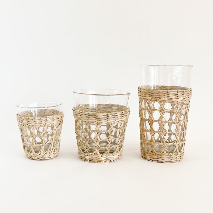 Seagrass cage glassware collection. Small tumbler, wide tumbler and highball glass shown. Each piece sold separately. Made from recycled glass and wrapped in a hand woven seagrass cage. Dishwasher safe when cage is removed.