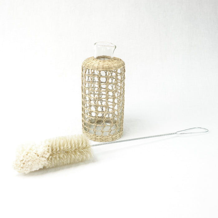 Natural bottle brush with wire handle laying next to a glass carafe wrapped in a seagrass cage. Each sold separately,