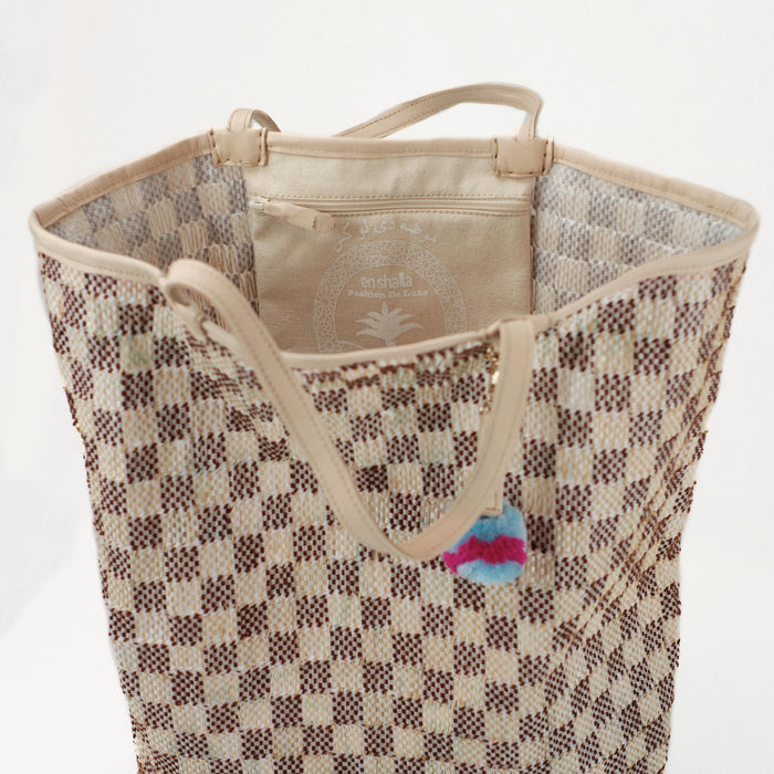 Interior view of brown and natural checkered beach tote with interior zip pocket. Natural lamb leather handles and trim with pale blue and hot pink pom-pom charm.