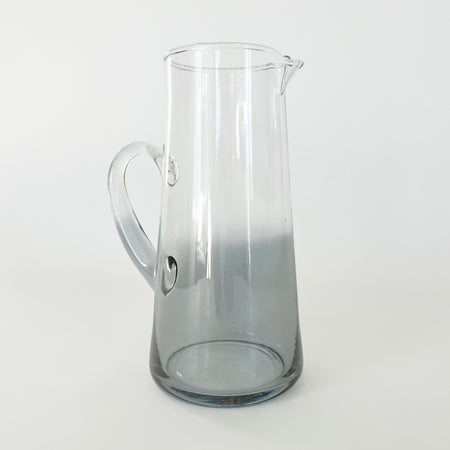 Smoke chroma glass pitcher has a soft coastal grey base that gently fades to clear glass near the top. Elegant single handle pitcher in modern silhouette. Measures 9.25" height, 4.5" diameter at base. 