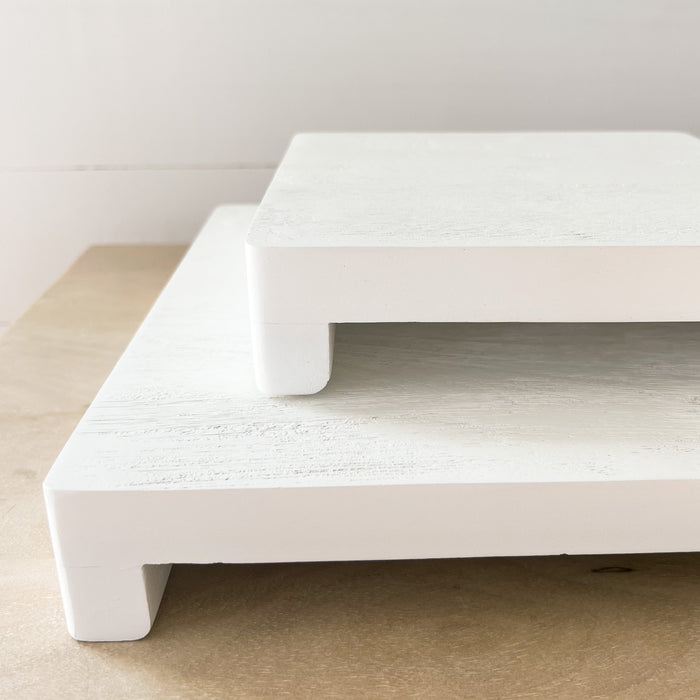 Small and large white wood serving boards/risers stacked together. Made of mango wood with a glossy white finish with natural grain showing though. Each sold separately.