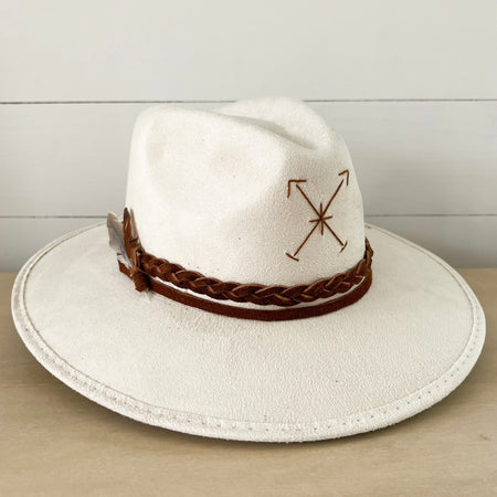 The Roads Hat in ivory suede. Crown is wrapped with sepia suede braided strap and finished with a small feather and crystal. Hand stitched arrows adorn the front of the hat. Measures 22" (58cm) at crown 