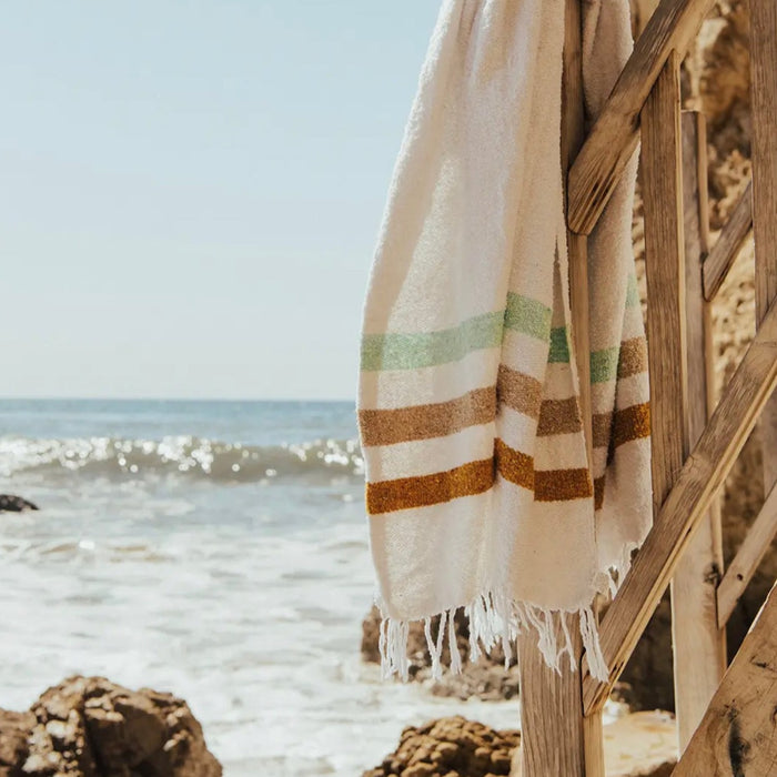 The Sol throw, woven from 100% post consumer materials in a blend of acrylic, polyester and cotton. Sustainability meets modern California style. White blanket bordered with graphic stripes in seafoam green, tan and rust. Measures 51" x 74".